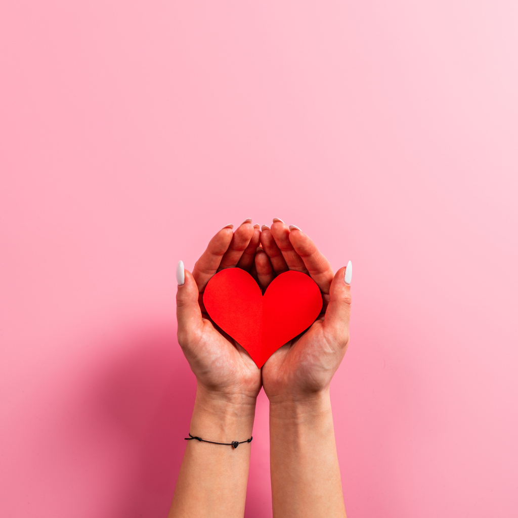 Красное бумажное сердце в руках на розовом фоне