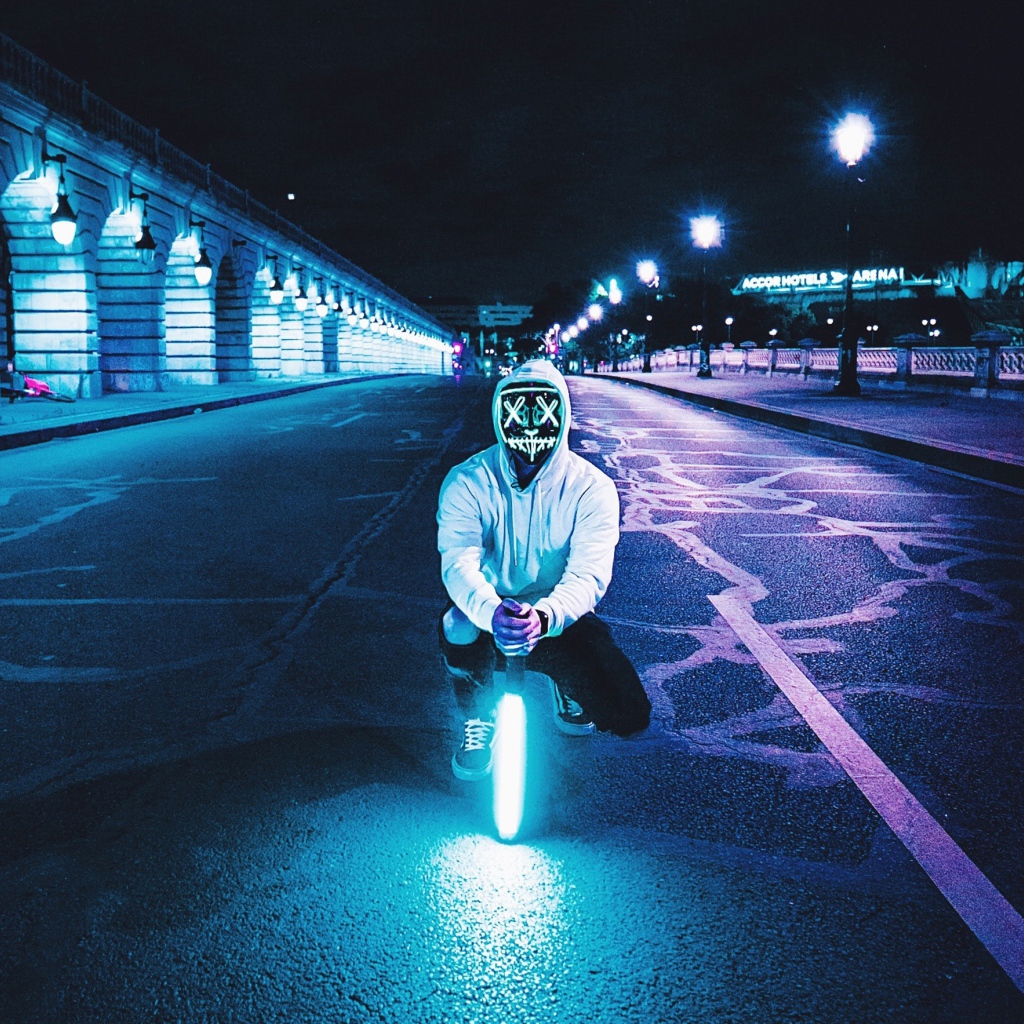 Парень в светящейся маске сидит с неоновой лампой на дороге