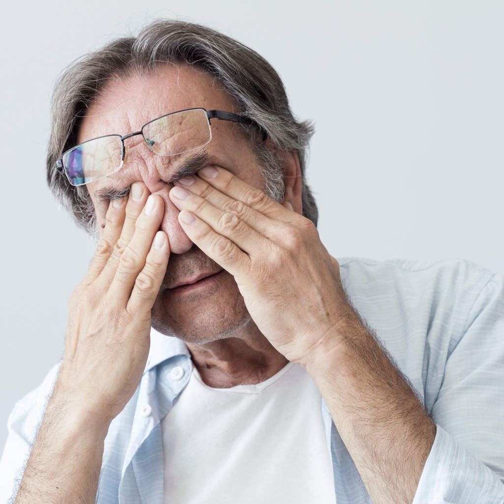 Пожилой мужчина трет глаза на сером фоне