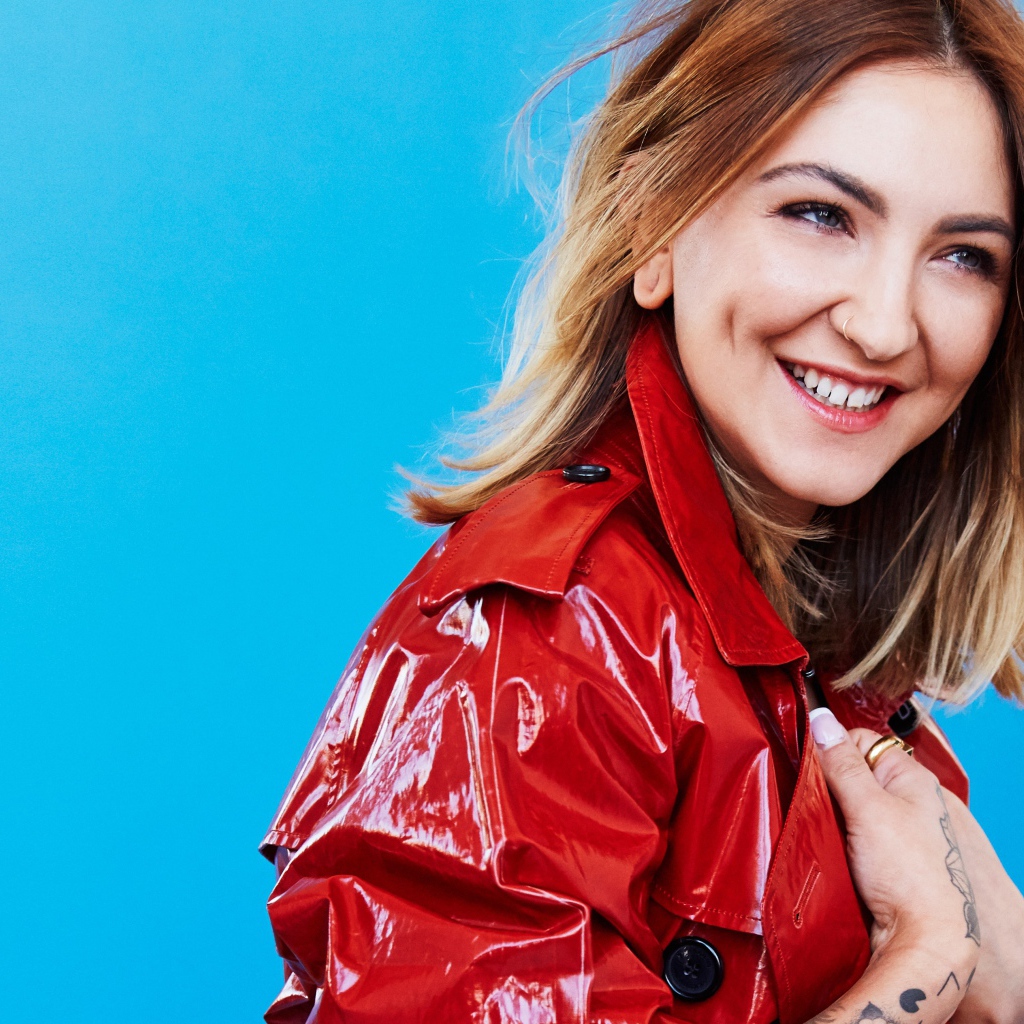 Улыбающаяся певица Джулия Майклз в красной куртке на голубом фоне