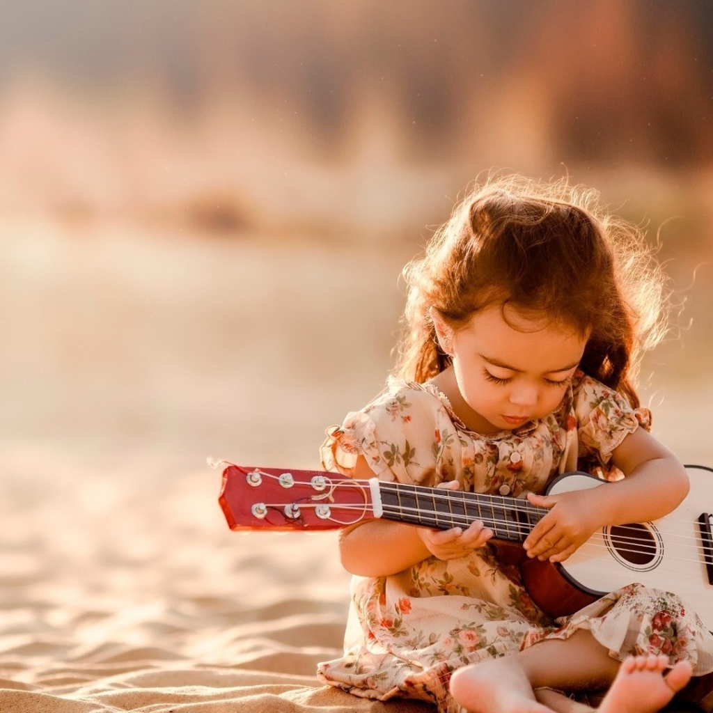 Маленькая девочка с гитарой сидит на песке 