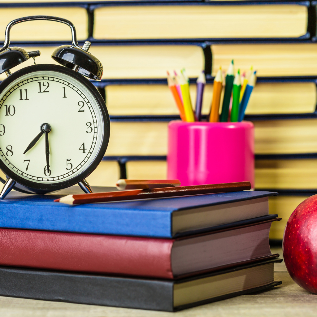 Будильник, книги, карандаши и яблоко на столе у стены 