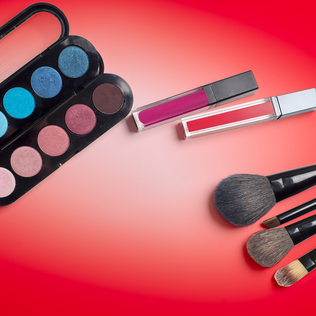 Блеск для губ, тени и кисточки на красном фоне
