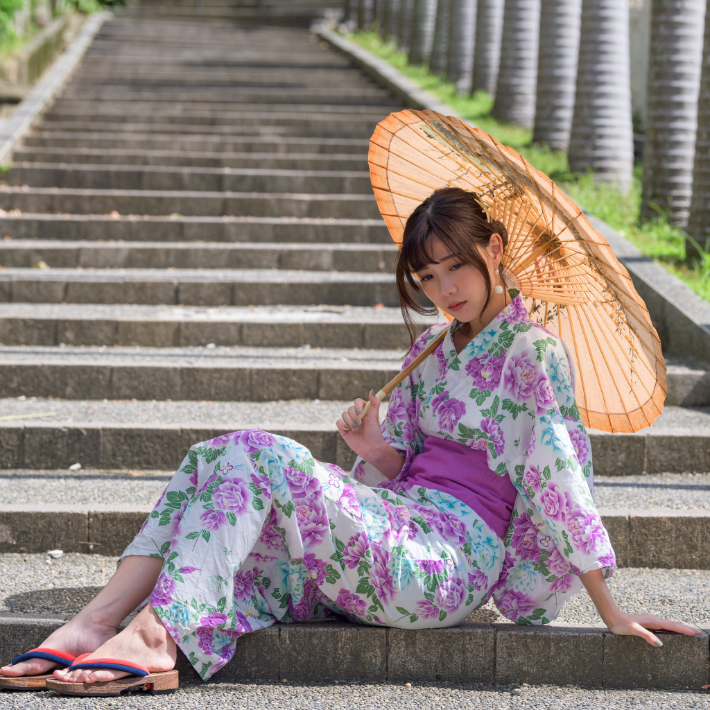 Девушка азиатка в кимоно с зонтом в руках сидит на ступеньках 