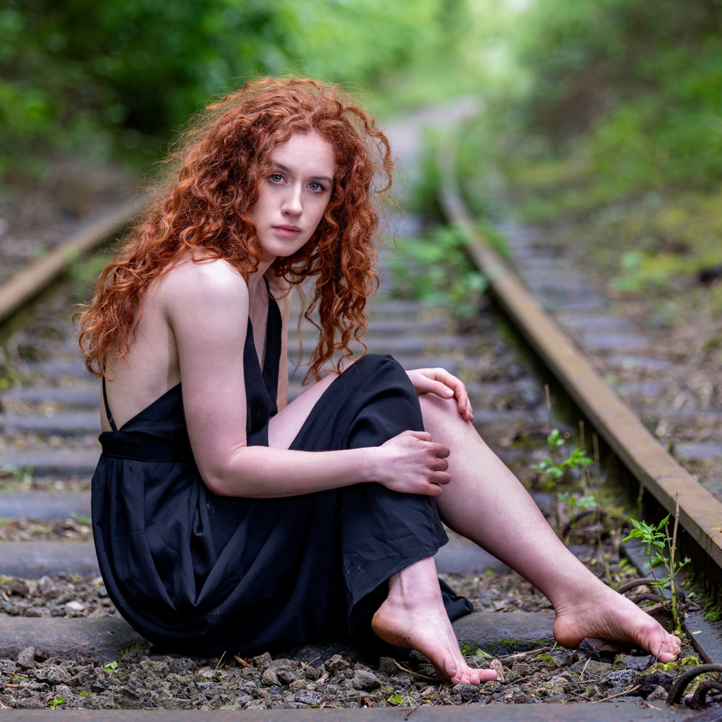 Рыжеволосая девушка в черном платье сидит на путях