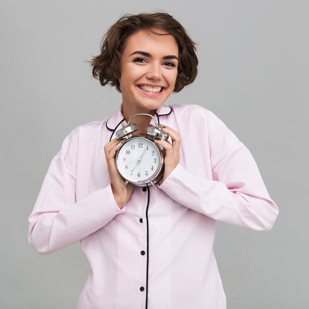 Улыбающаяся девушка в розовой рубашке с будильником в руках 