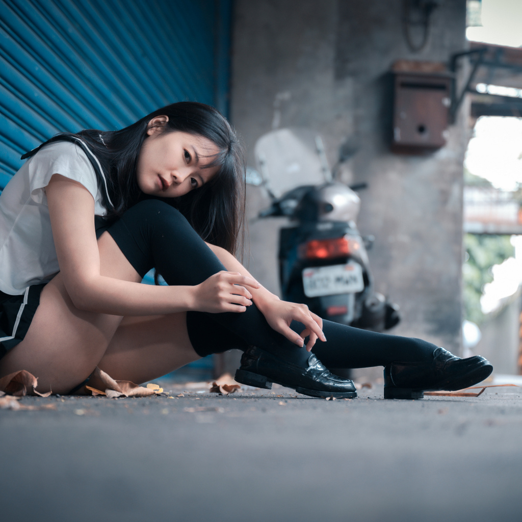 Молодая азиатка в черных чулках сидит у стены 