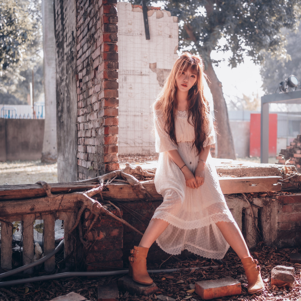 Молодая девушка азиатка в белом платье сидит на разваленной стене