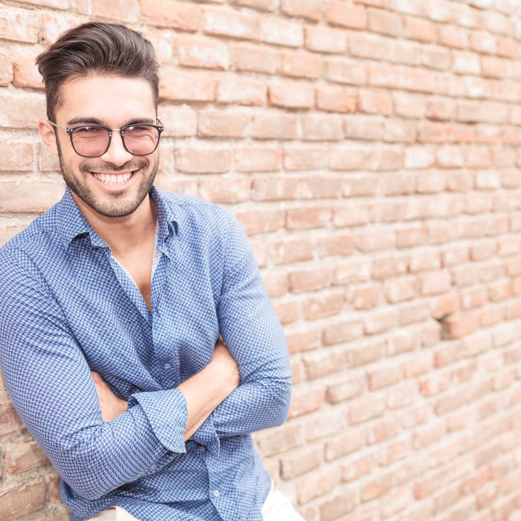 Улыбающийся мужчина в очках стоит у стены 