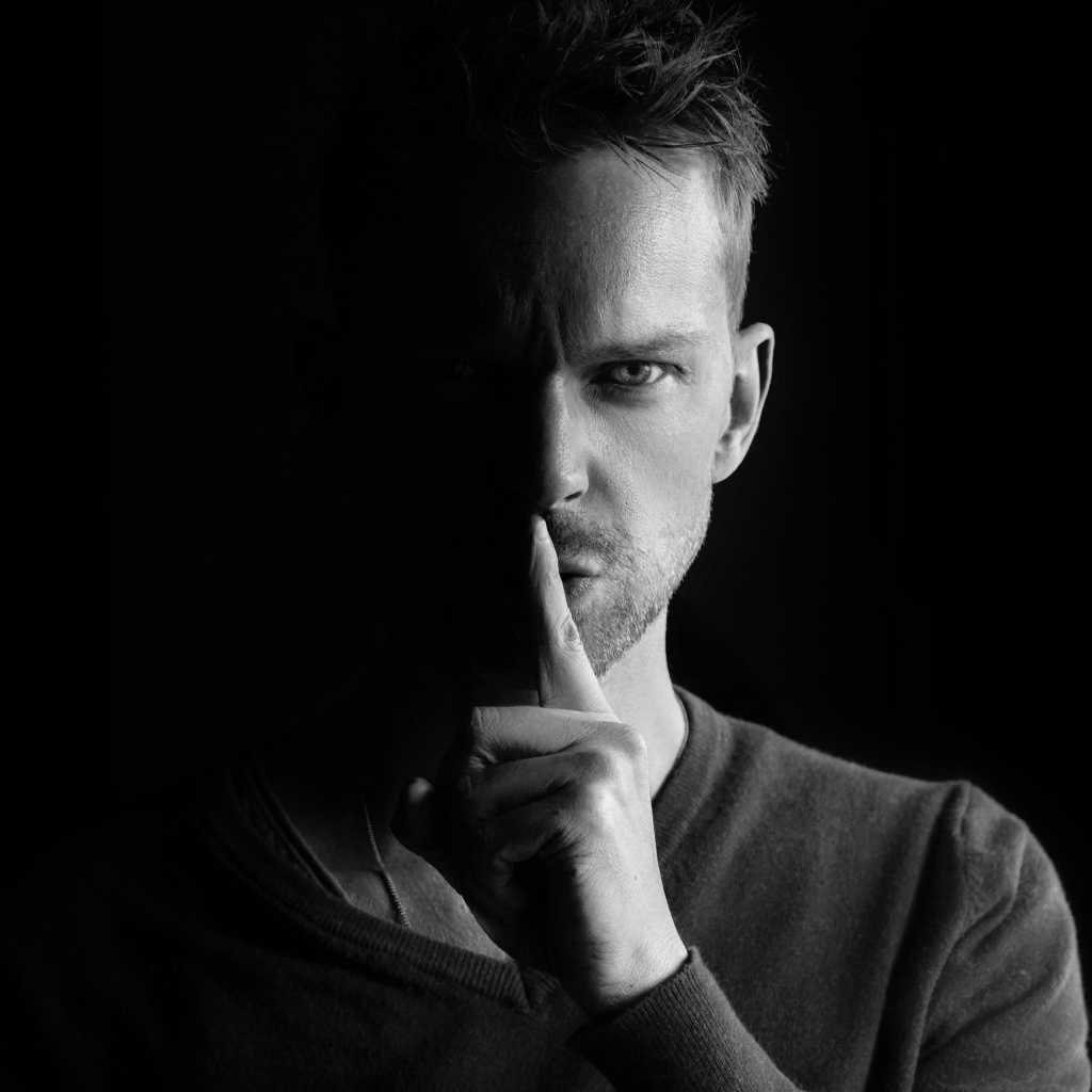 Молодой мужчина закрыл рот пальцем на черном фоне 