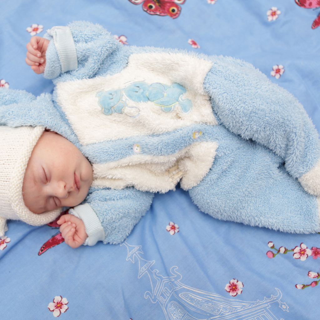 Грудной ребенок в голубом костюме лежит на кровати 