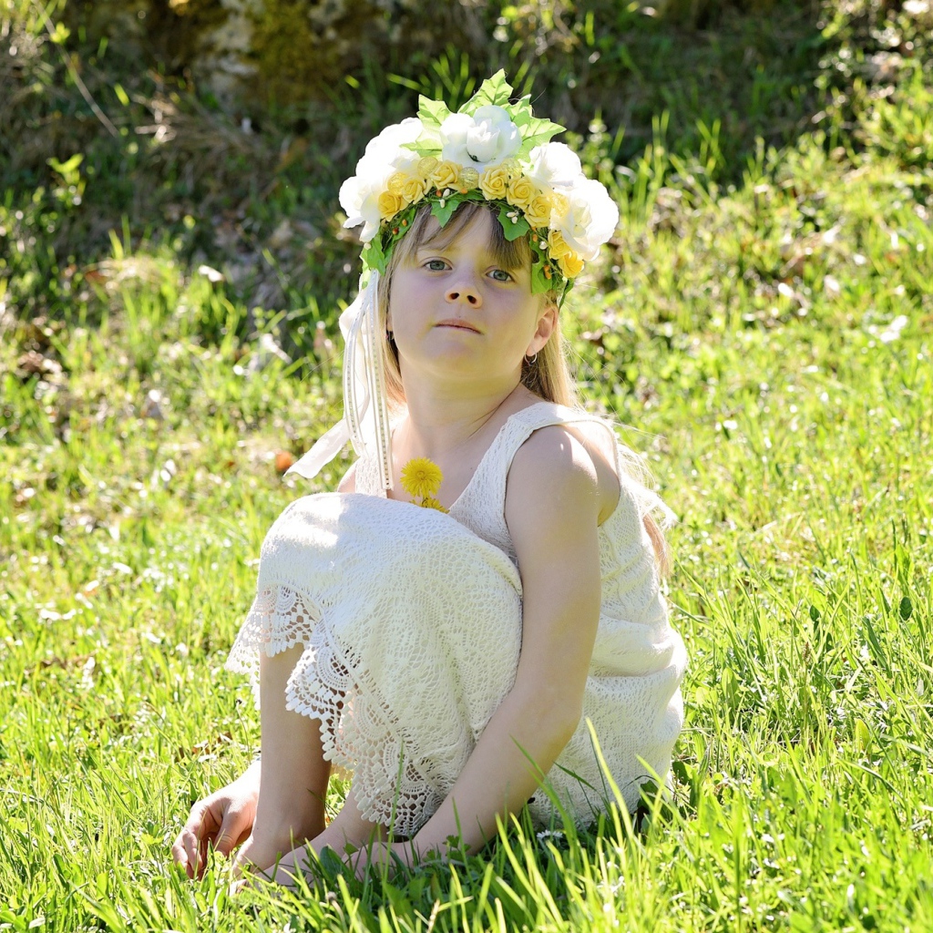 Маленькая девочка с венком на голове сидит на зеленой траве