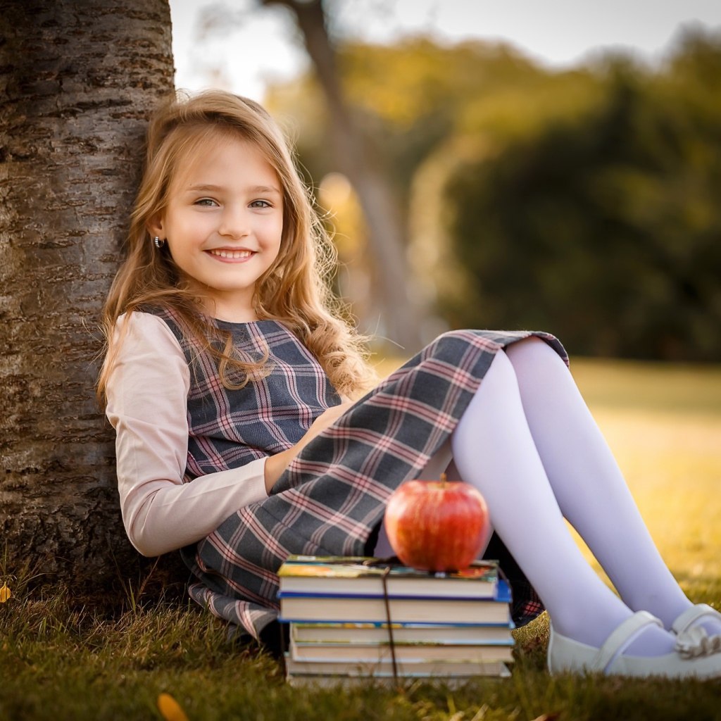 Маленькая девочка с книгами и яблоком сидит под деревом 