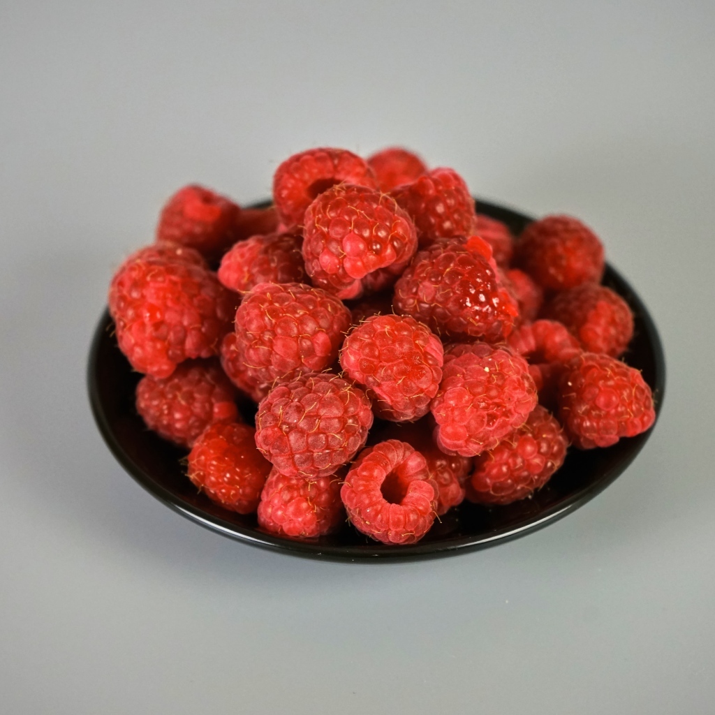 Спелая красная малина в черной тарелке на сером фоне