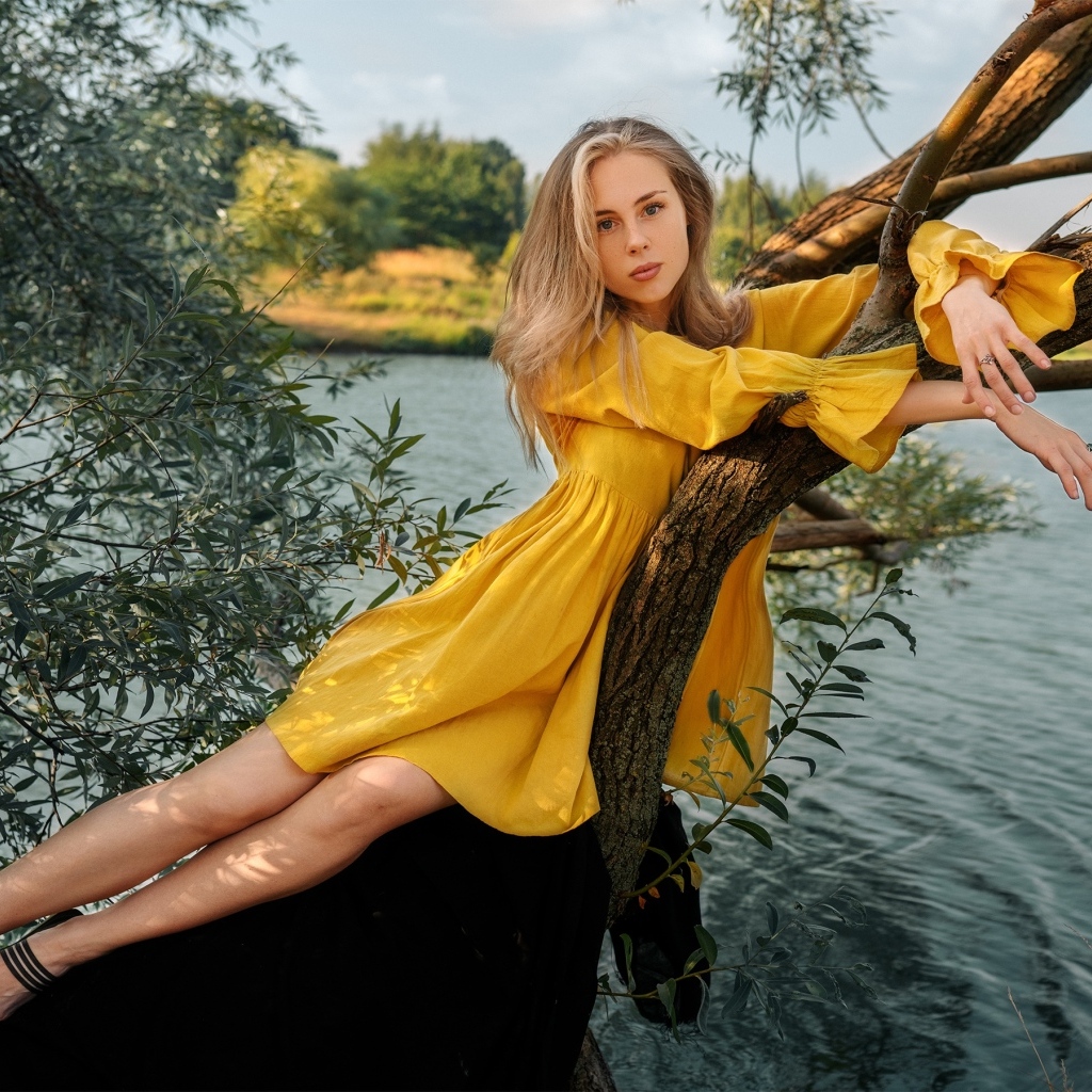 Красивая девушка в желтом платье лежит на дереве у воды