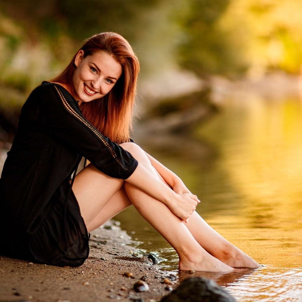 Улыбающаяся девушка в черном платье сидит у воды