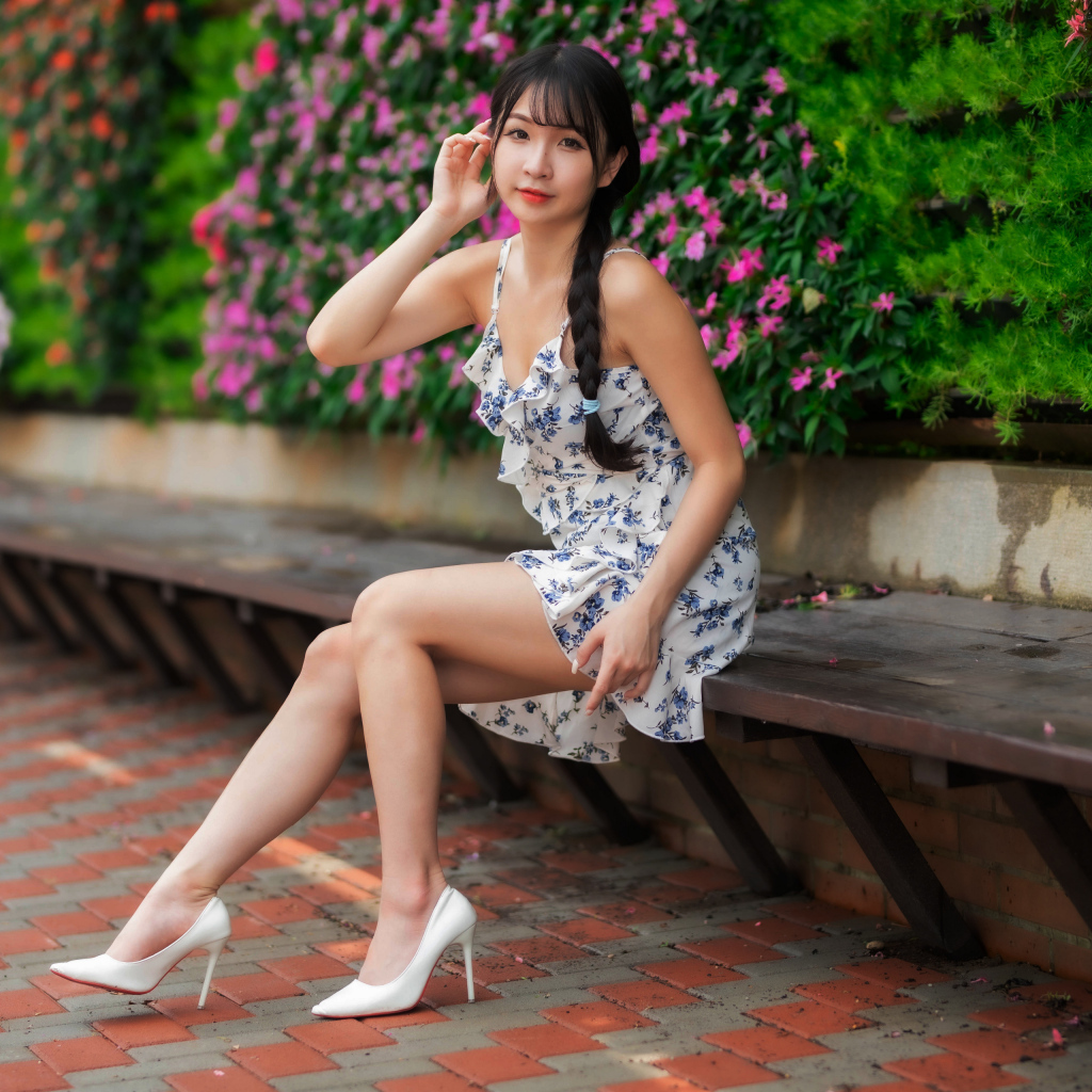 Азиатка в платье сидит на лавке у цветущих растений