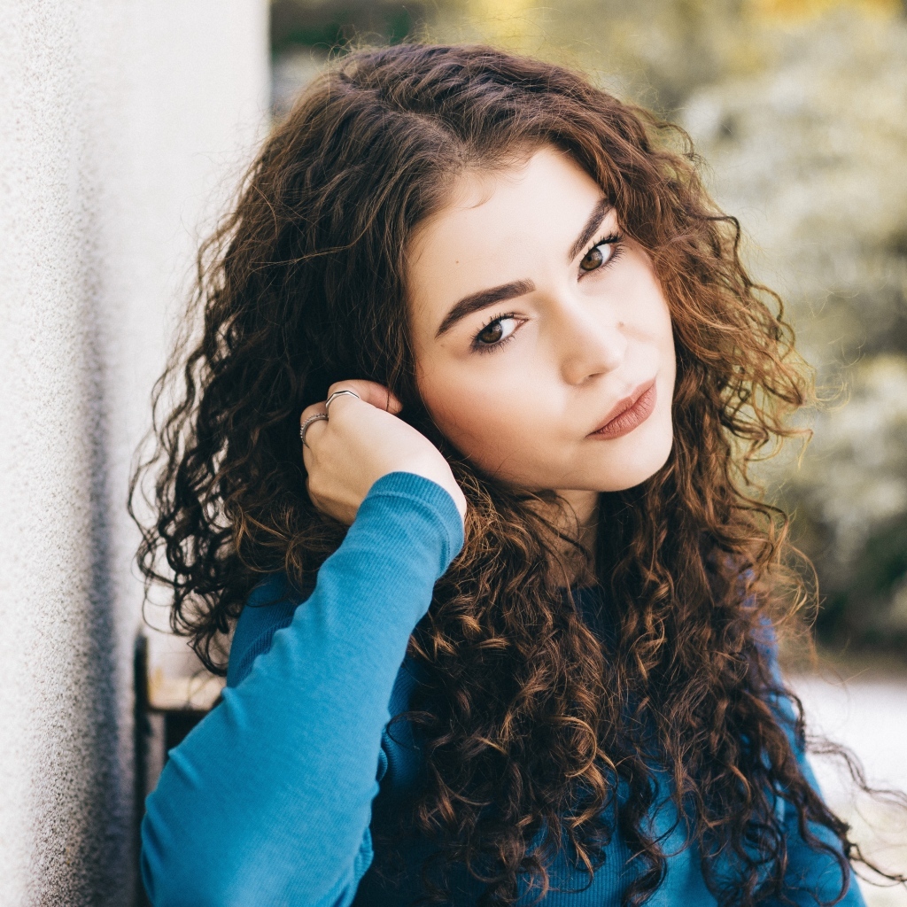 Кареглазая девушка с кудрявыми волосами стоит у стены