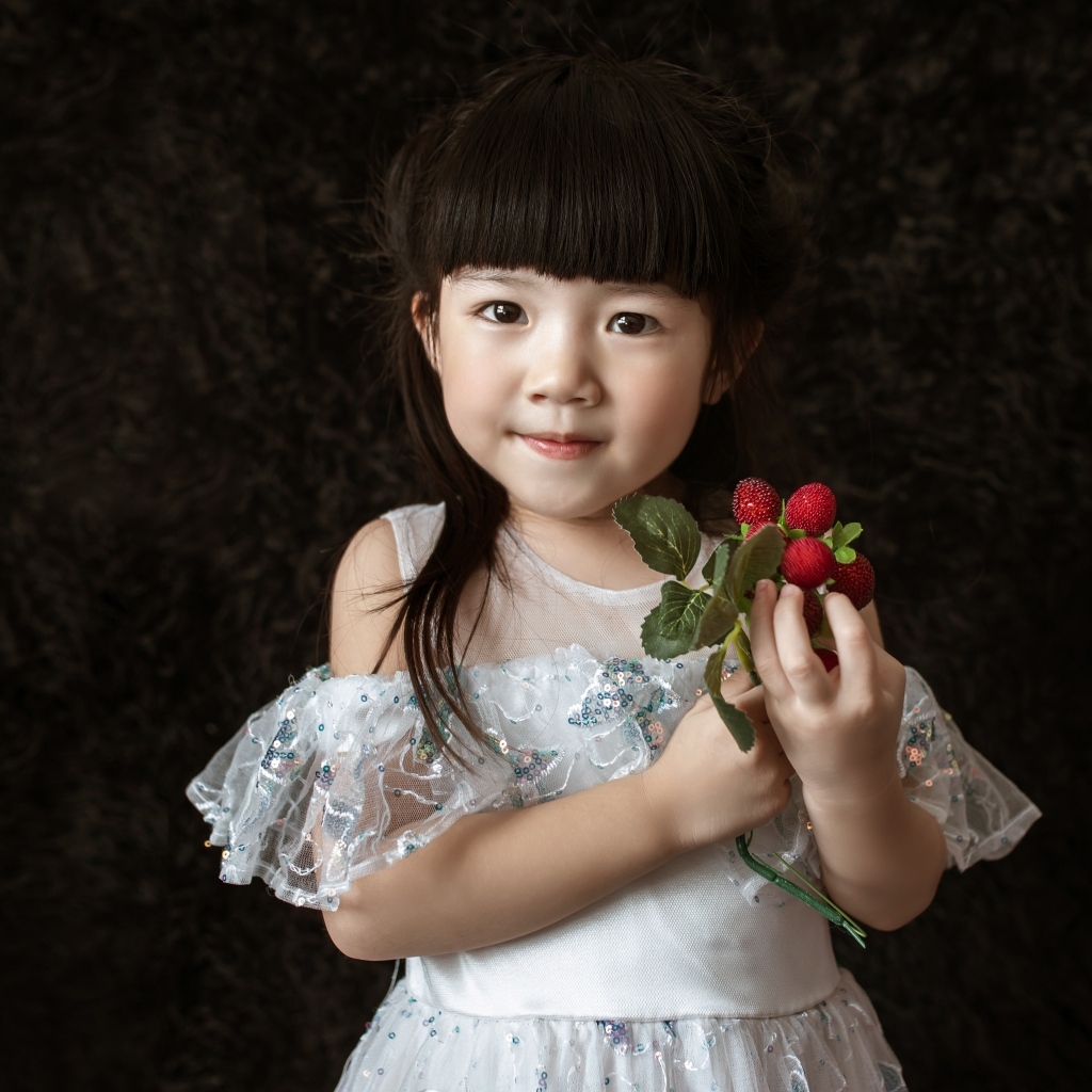 Маленькая девочка азиатка в белом платье с ягодами в руке на черном фоне