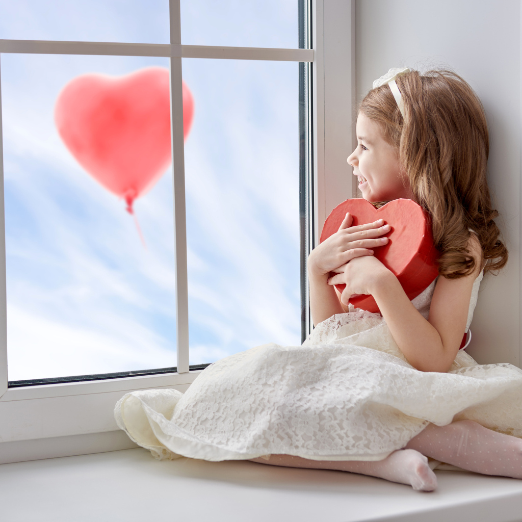 Маленькая девочка с коробкой в форме сердца сидит у окна