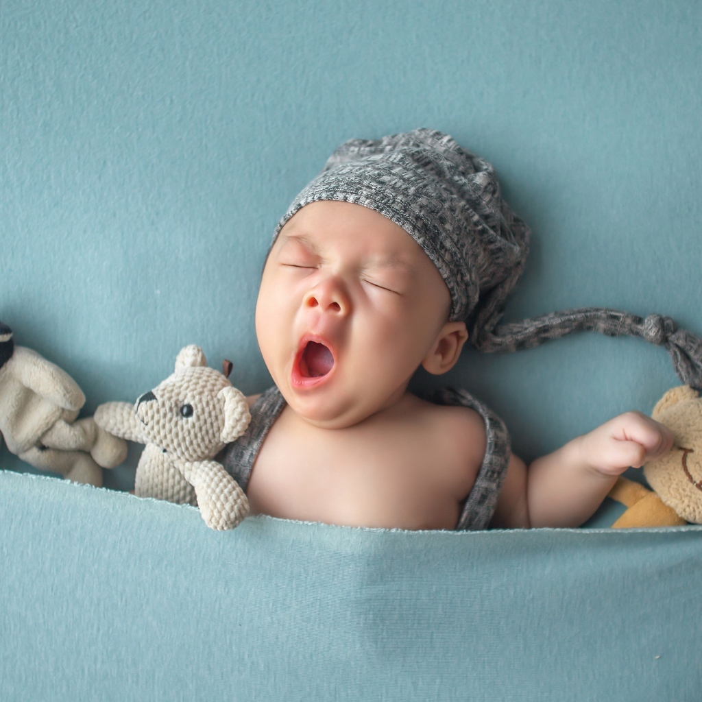 Зевающий малыш с игрушками под серым покрывалом