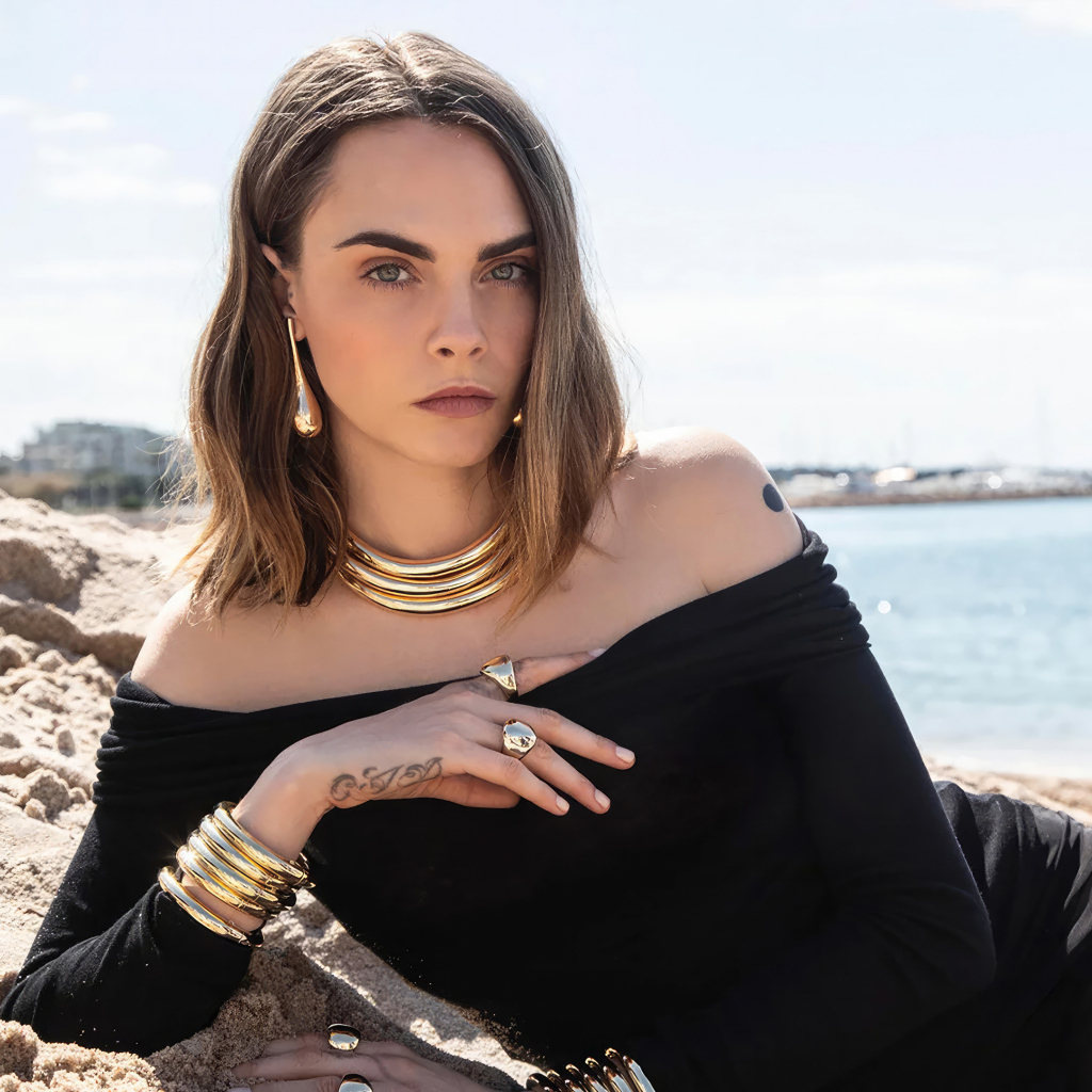 Кара Делевинь в черном платье лежит на песке