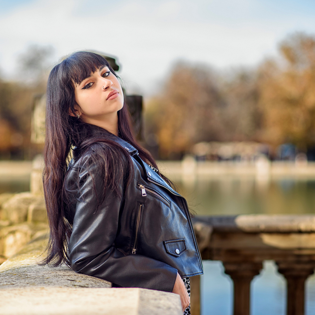 Девушка в черной куртке стоит на мосту у воды