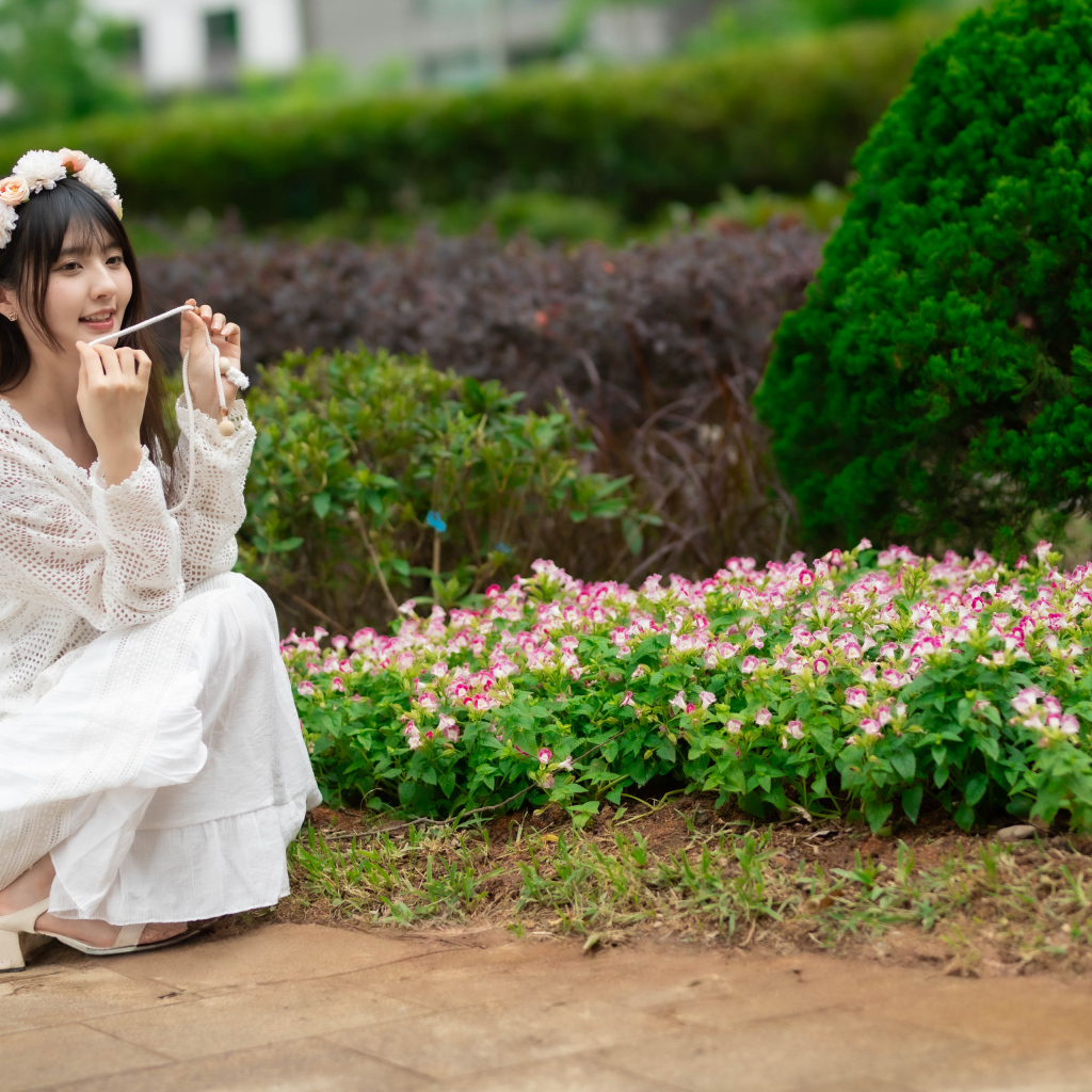 Милая азиатка в белом наряде сидит у клумбы с цветами