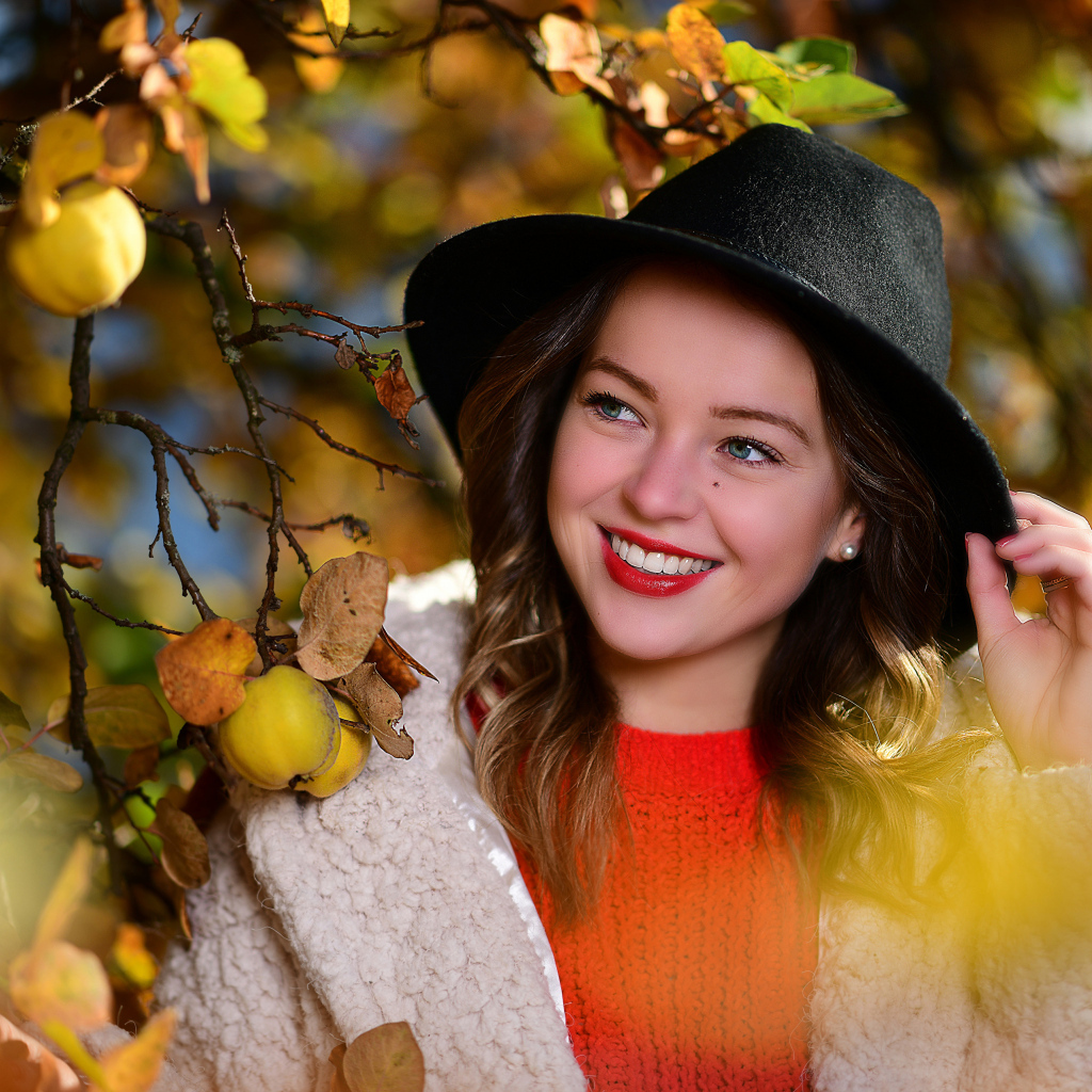Улыбающаяся девушка в черной шляпе у дерева с яблоками