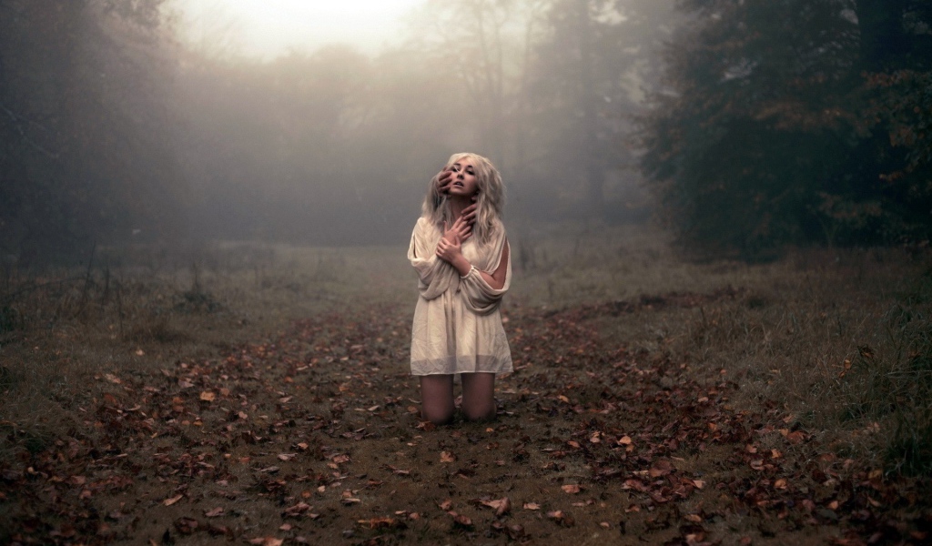 Блондинка в белом платье стоит на коленях среди леса