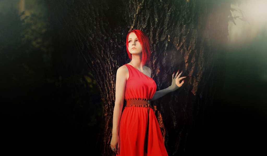 Рыжая девушка в красном платье стоит у дерева
