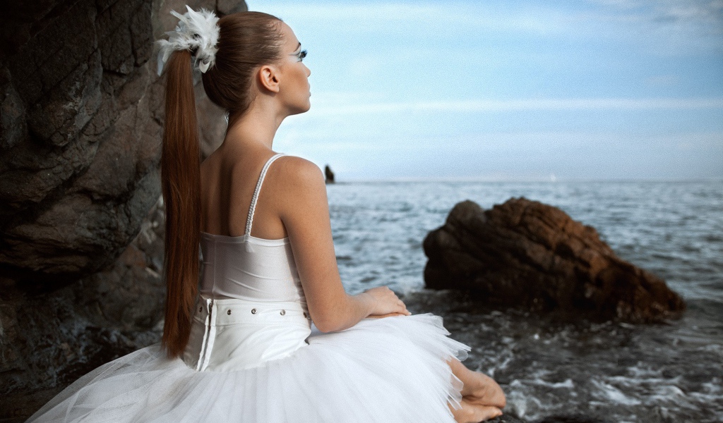 Балерина в белом платье сидит на берегу моря