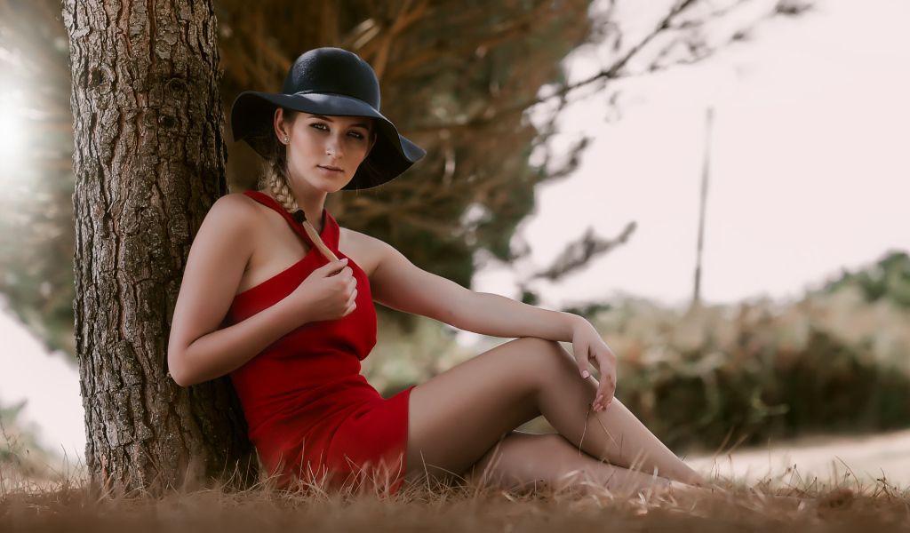 Красивая девушка в красном платье и черной шляпе сидит под деревом