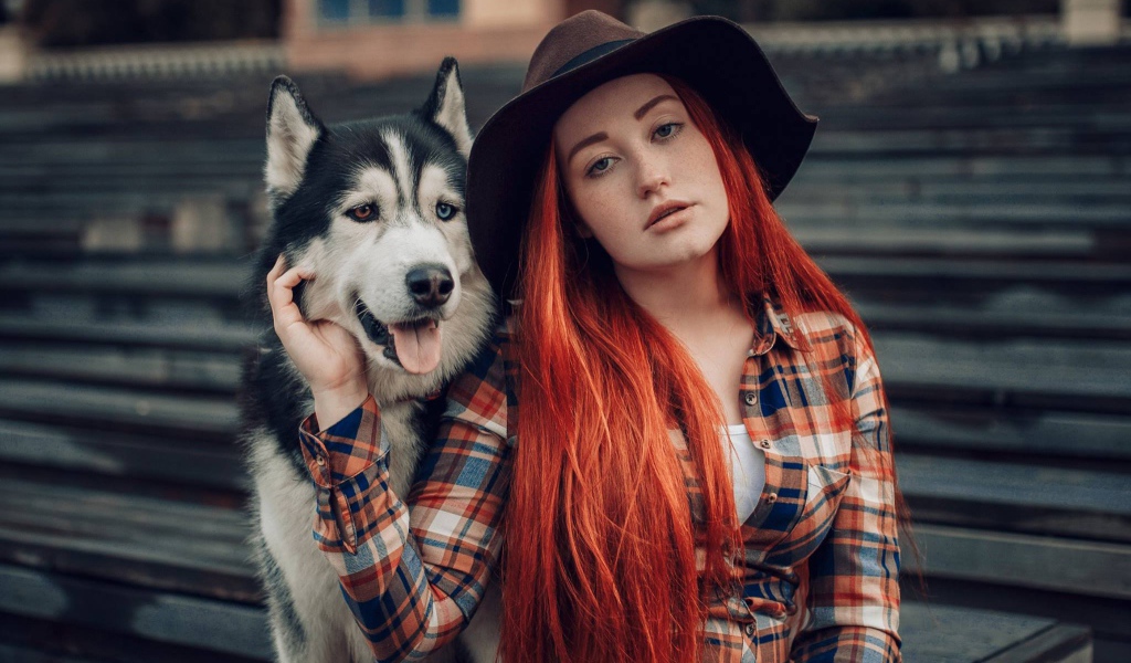 Рыжеволосая девушка в черной шляпе сидит с хаски