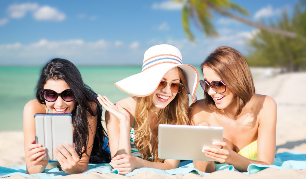 Три улыбающиеся девушки загорают на пляже с планшетами в руках