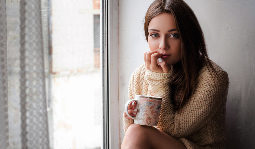 Молодая девушка в свитере сидит с чашкой на подоконнике