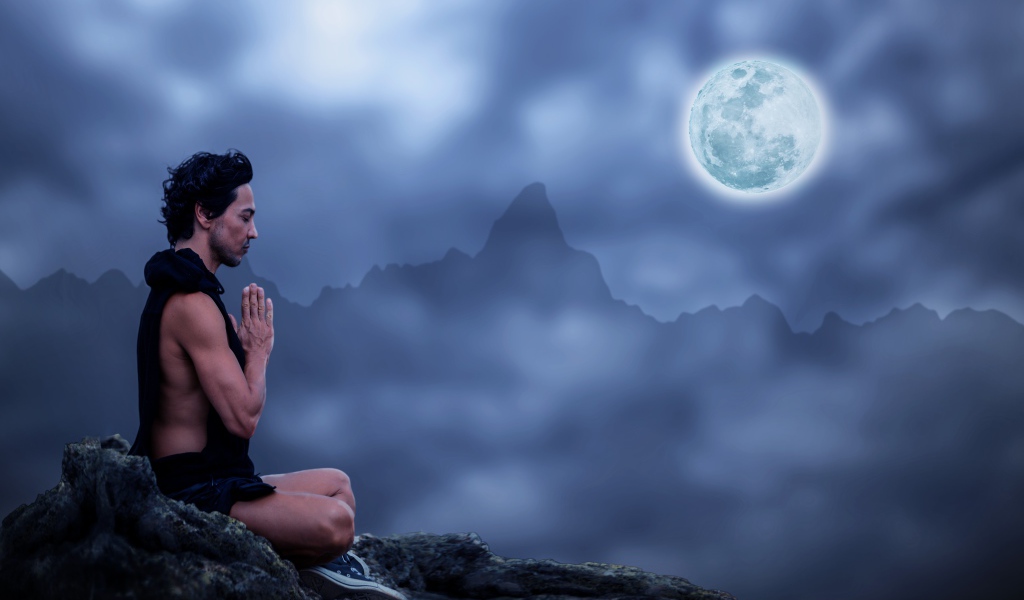 Мужчина медитирует на камне ночью в полнолуние