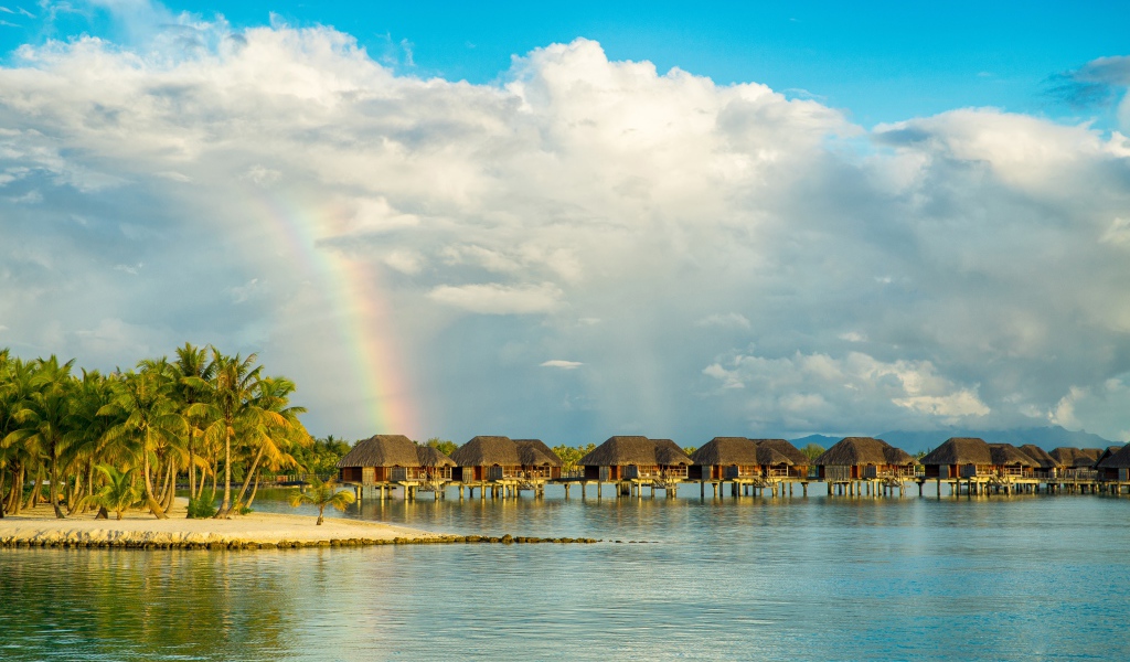Бунгало в воде на тропическом пляже под облачным небом с радугой