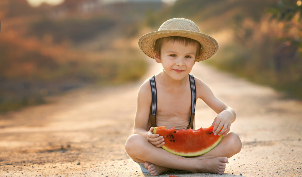 Маленький мальчик в шляпе сидит на дороге с долькой арбуза