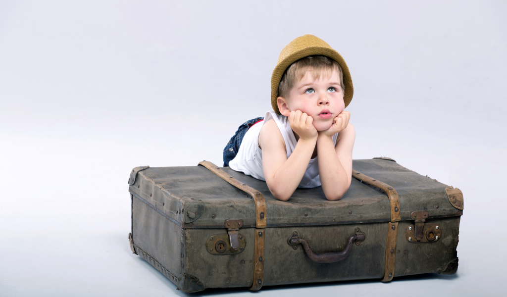 Маленький мальчик лежит на большом чемодане на сером фоне