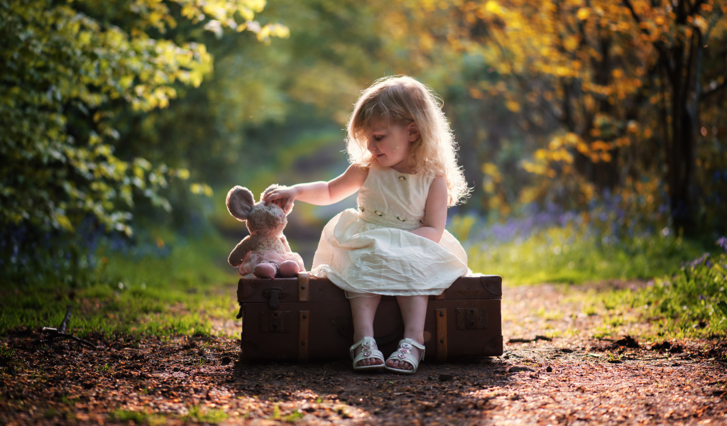 Маленькая девочка в белом платье сидит на большом чемодане с игрушкой
