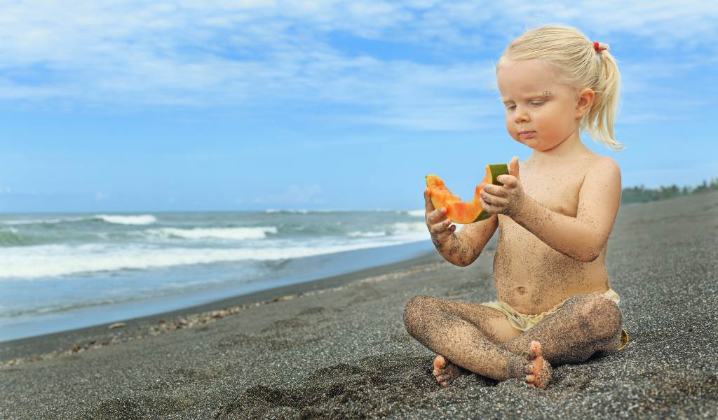 Маленькая девочка сидит на песке у моря с кусочком дыни