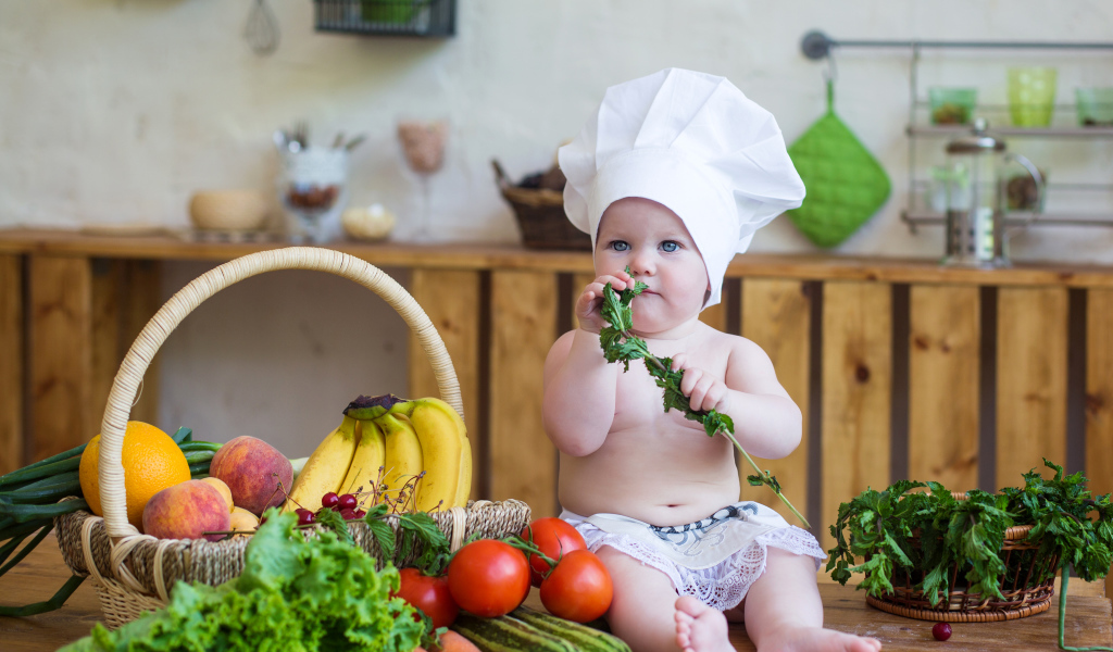 Маленький ребенок в шапке повара сидит на столе с овощами