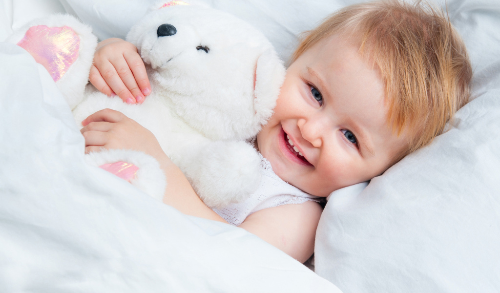 Улыбающийся ребенок лежит в кровати с белым плюшевым медвежонком