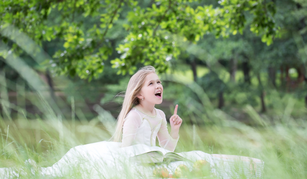 Красивая маленькая девочка в белом платье с книгой сидит на траве