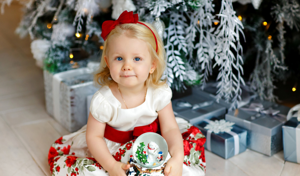 Маленькая девочка блондинка сидит с подарками под новогодней елкой