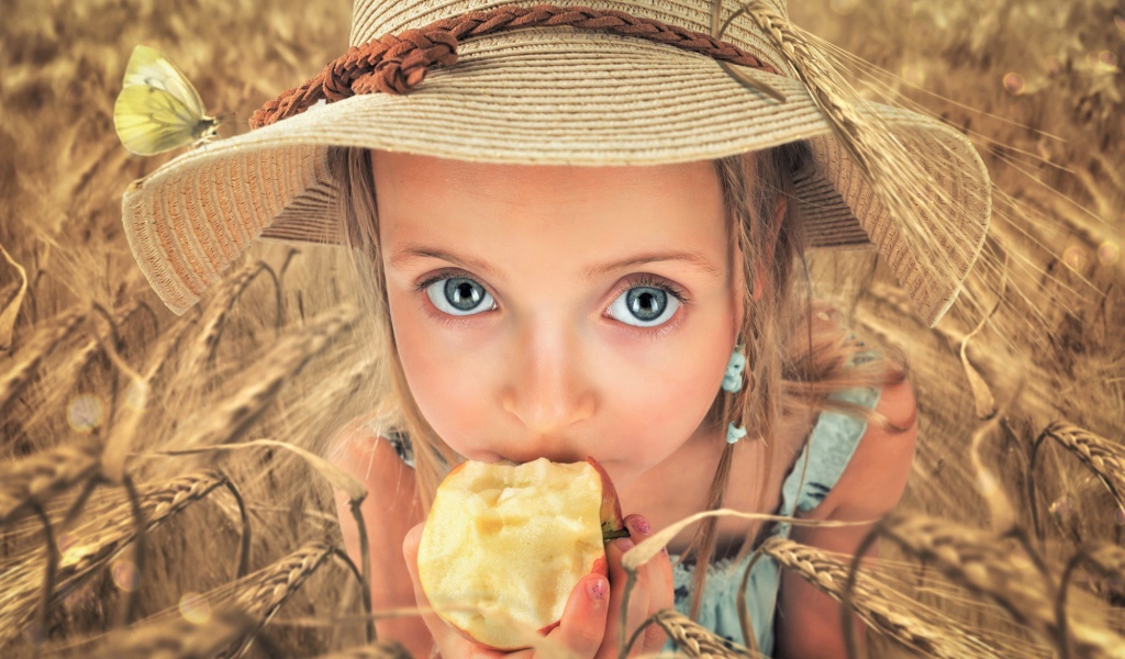 Маленькая голубоглазая девочка в шляпе ест яблоко