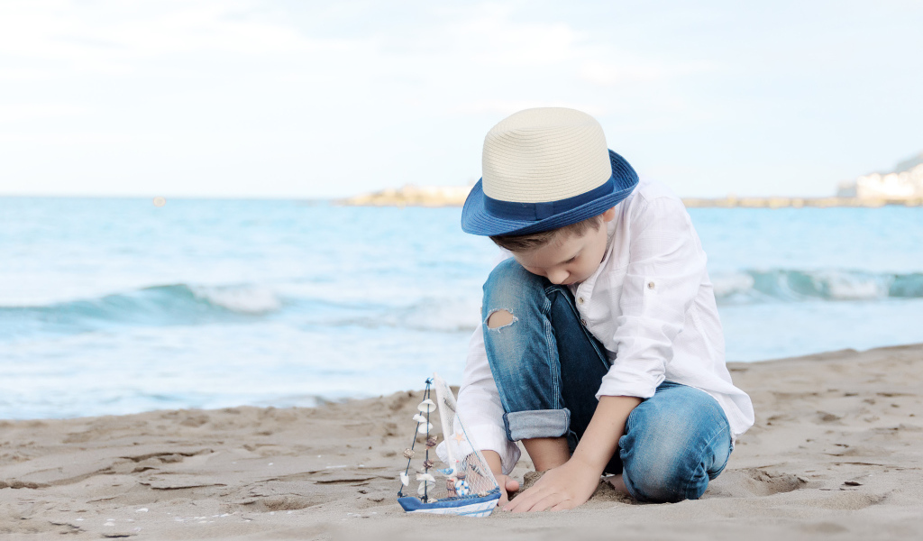 Маленький мальчик в шляпе играет с игрушечным корабликом на песке