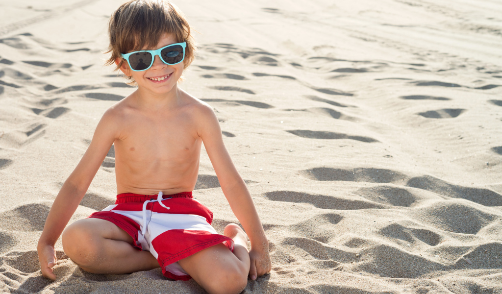 Маленький мальчик в очках сидит на песке на пляже