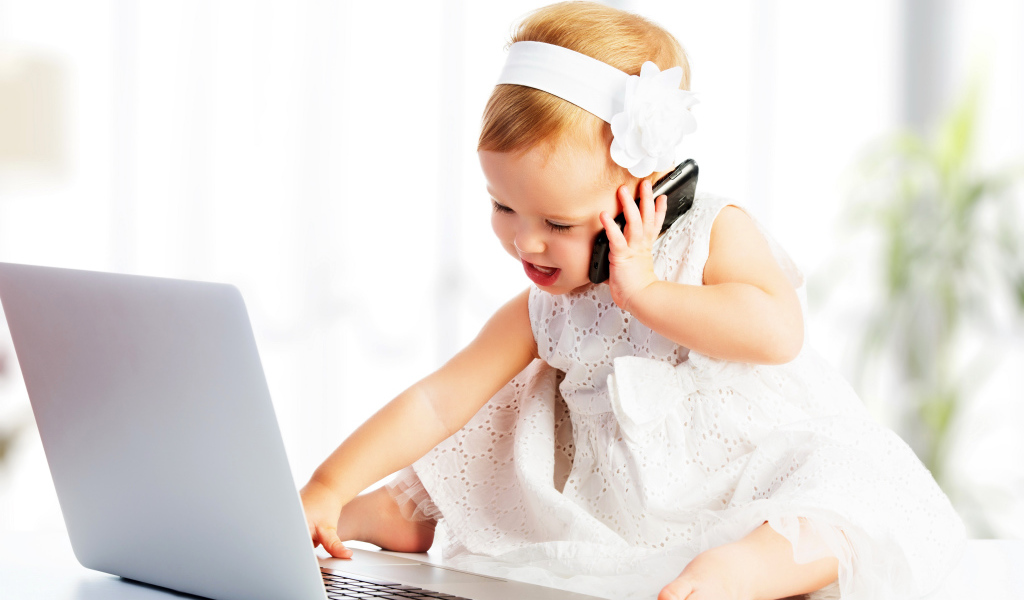 Маленькая девочка в белом платье сидит с телефоном у ноутбука